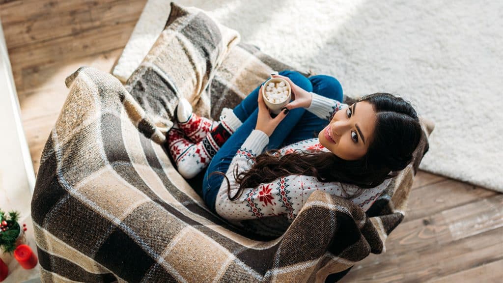 top view of young woman enjoying hot chocolate with marshmallow while sitting in cozy armchair