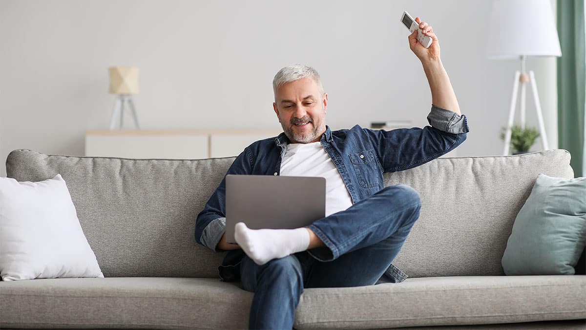 Happy senior man enjoying time at home, surfing on Internet, using notebook, holding remote for air conditioner